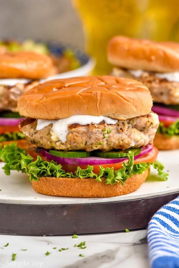 Photo of Greek Turkey Burgers served on a bun with yogurt sauce, lettuce, tomato, pickles, and onions.
