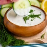 Pinterest graphic for Yogurt Sauce recipe. Photo shows person's hand dipping a slice of cucumber into a bowl of Yogurt Sauce. Text says, "Yogurt Sauce simplejoy.com"