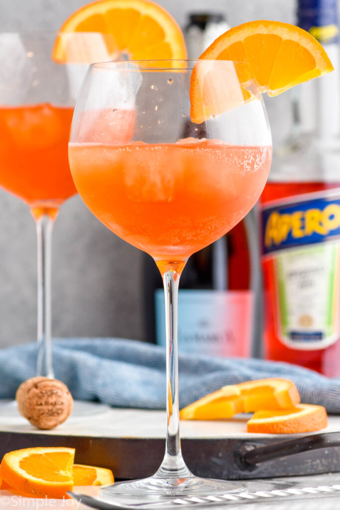 Close up photo of Aperol Spritz cocktails garnished with orange slices. Bottles of sparkling wine and aperol in the background.