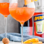 Close up photo of Aperol Spritz garnished with orange slices. Bottles of sparkling wine and aperol in the background.