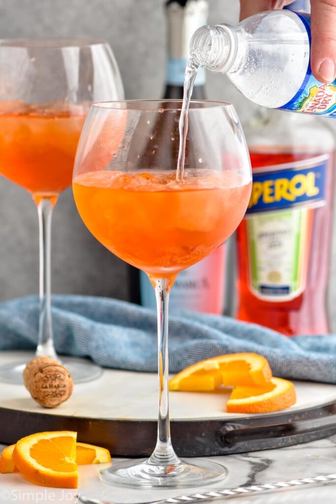Photo of person's hand pouring club soda into glass for Aperol Spritz recipe. Bottles of sparkling wine and aperol in the background, along with orange slices for garnish.