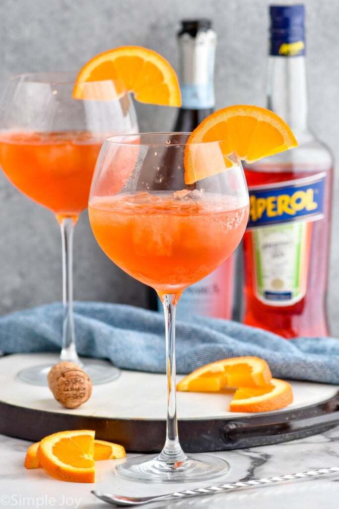 Photo of Aperol Spritz cocktails garnished with orange slices. Bottles of sparkling wine and aperol in the background.