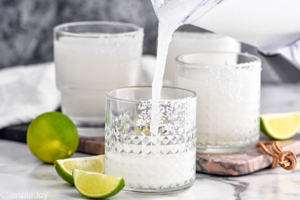 Close up photo of a blender of Frozen Margarita recipe being poured into glasses with salted rims. Lime slices beside glasses for garnish.