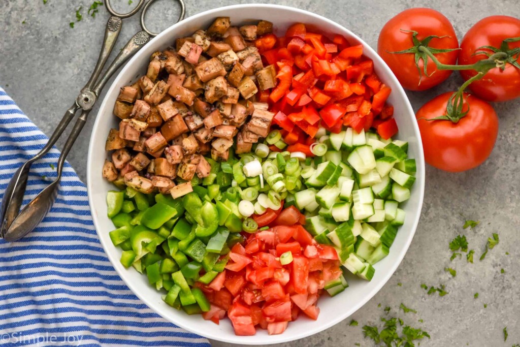 Overhead photo of a bowl of ingredients for Eggplant Salad recipe. Tomatoes and serving tongs beside bowl.
