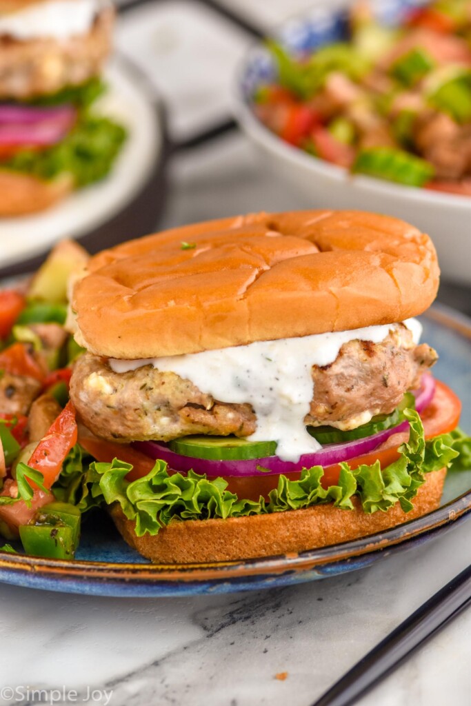 Close up photo of Greek Turkey Burgers served on buns with yogurt sauce, lettuce, tomato, onion, and pickles.