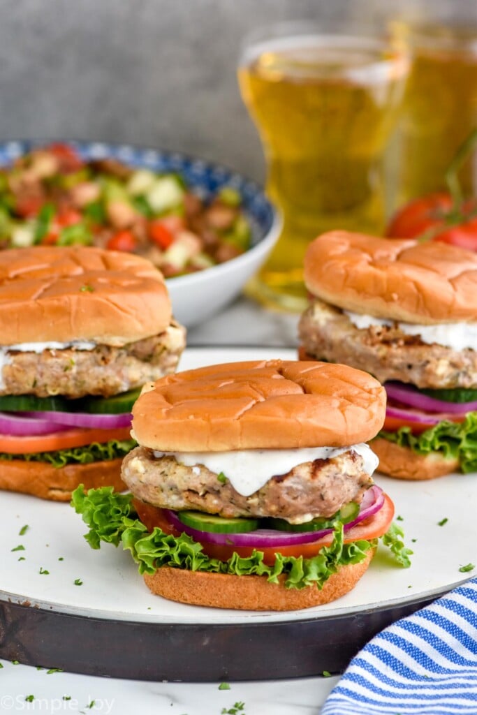 Close up photo of Greek Turkey Burgers served on buns with yogurt sauce, lettuce, tomato, onion, and pickles. Two glasses of beer and bowl of eggplant salad in the background.