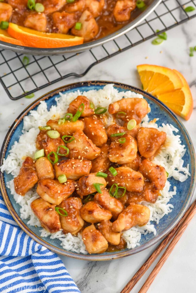 Overhead photo of Orange Chicken recipe served on a plate of rice. Slices of orange and chopsticks beside plate.