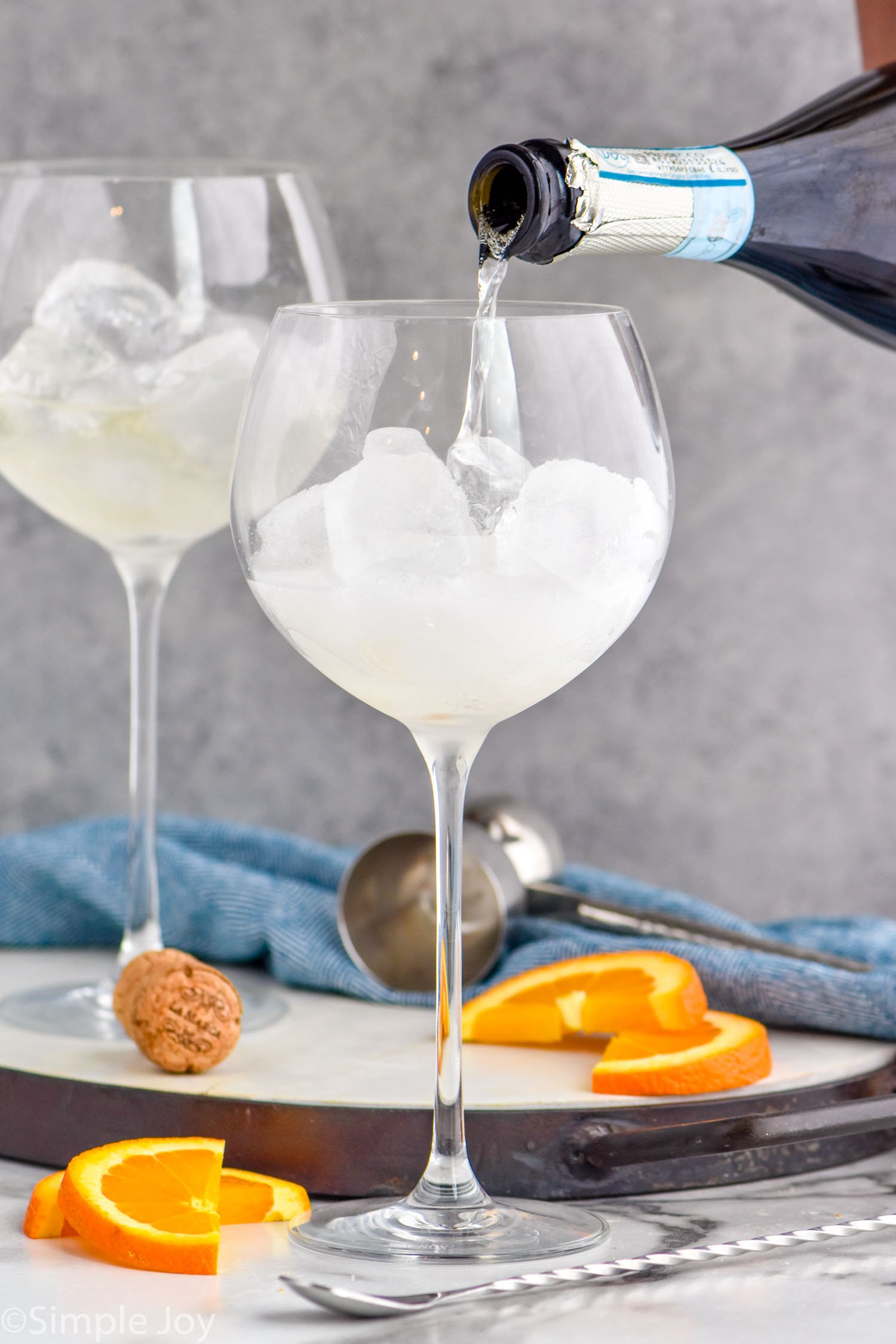 Photo of sparkling wine being poured into glass of ice for Aperol Spritz recipe. Orange slices on counter for garnish.