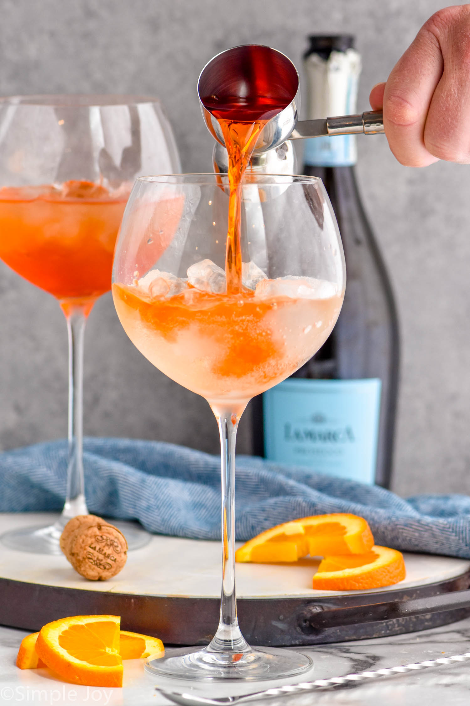 Photo of person's hand pouring ingredients for Aperol Spritz recipe into a glass. Bottle of sparkling wine and orange slices for garnish beside glasses