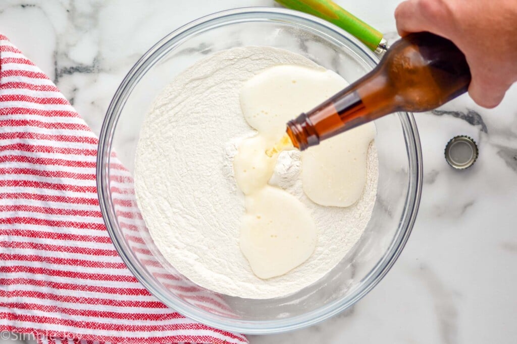 Overhead photo of a mixing bowl of ingredients for Beer Bread recipe. Person's hand is pouring beer into bowl.