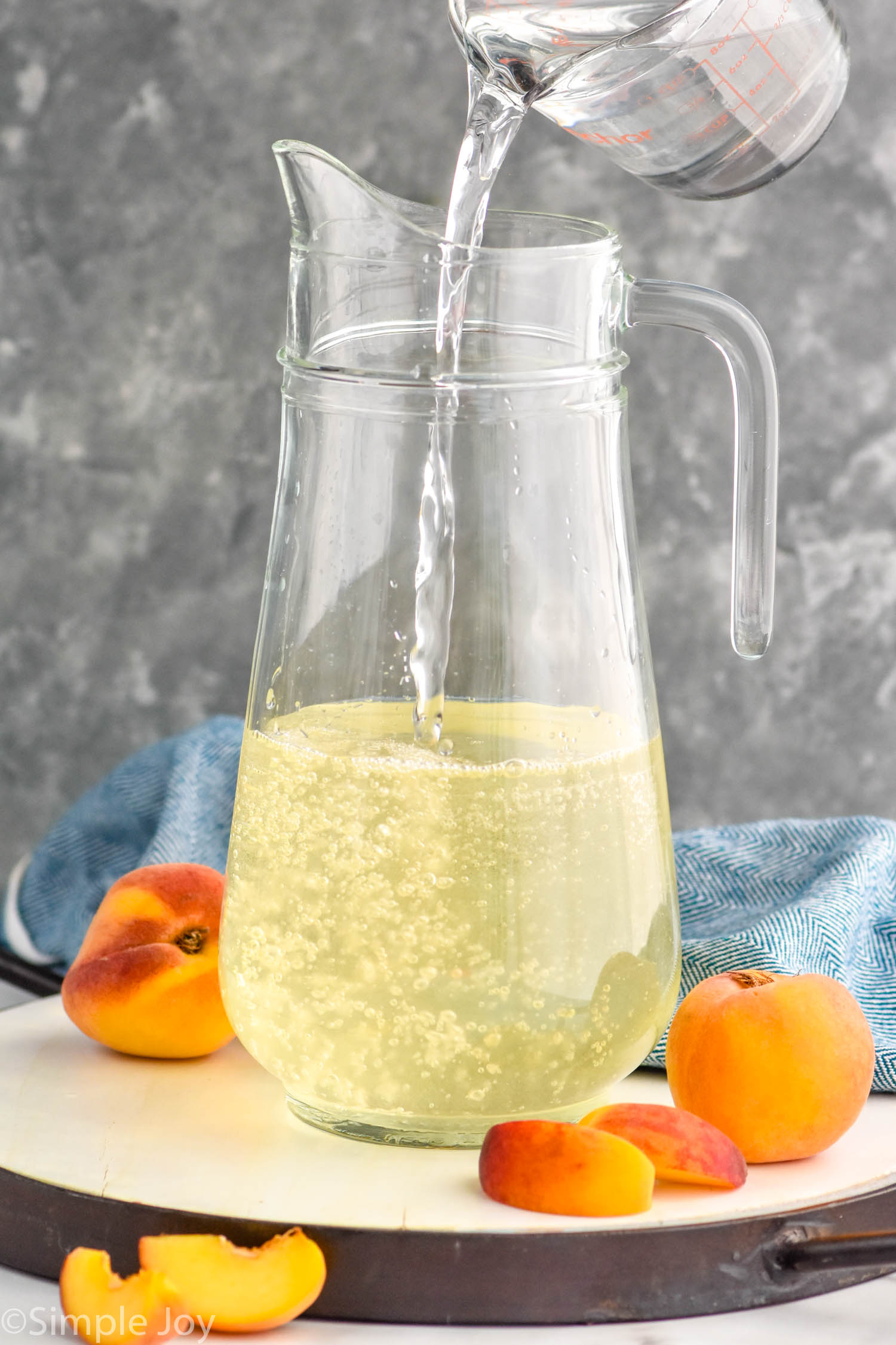 Photo of measuring cup of ingredients being poured into pitcher for Peach Sangria recipe. Peaches beside pitcher.