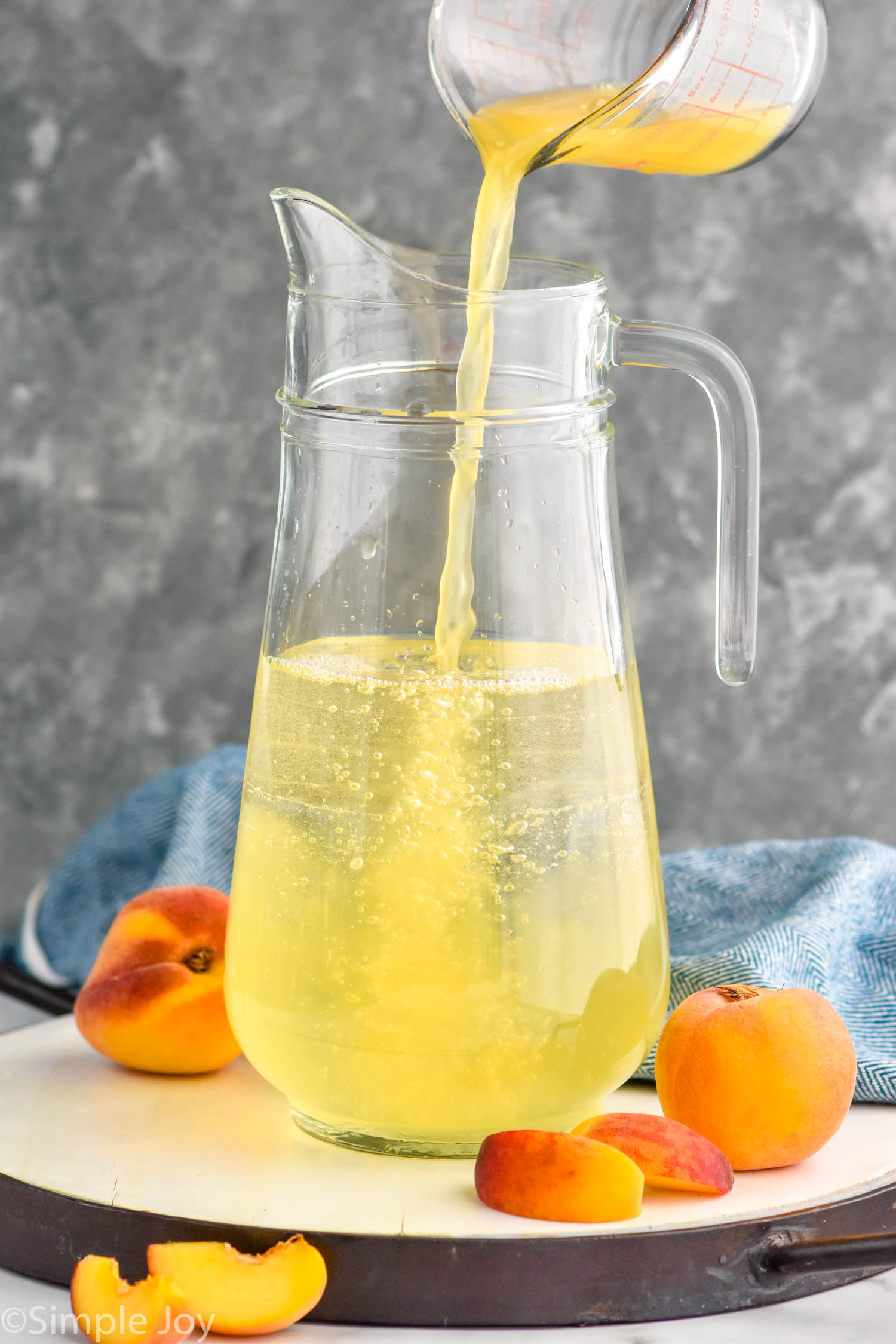 Photo of measuring cup of ingredients being poured into pitcher for Peach Sangria recipe. Peaches beside pitcher.