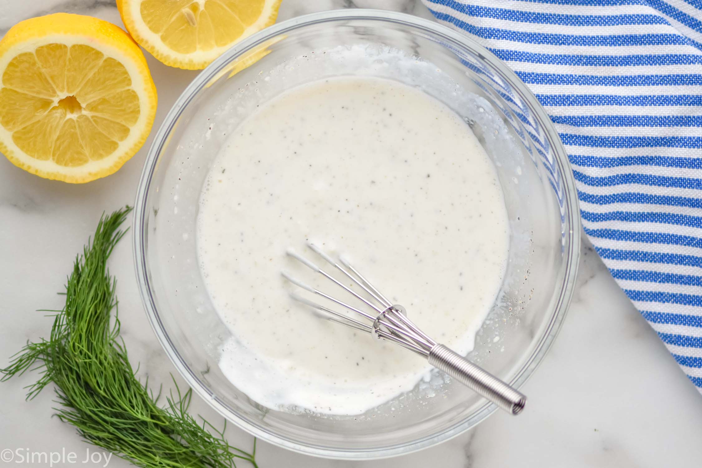 Overhead photo of a mixing bowl of Yogurt Sauce with a whisk. Lemons, dill, and dish towel sit beside bowl.