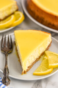 Slice of Lemon Cheesecake served on a plate with a fork and two lemon slices.