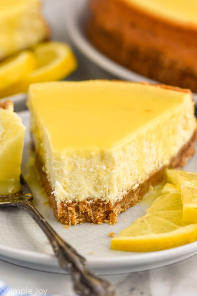 Close up photo of a slice of Lemon Cheesecake served on a plate with a fork and garnished with two lemon slices.