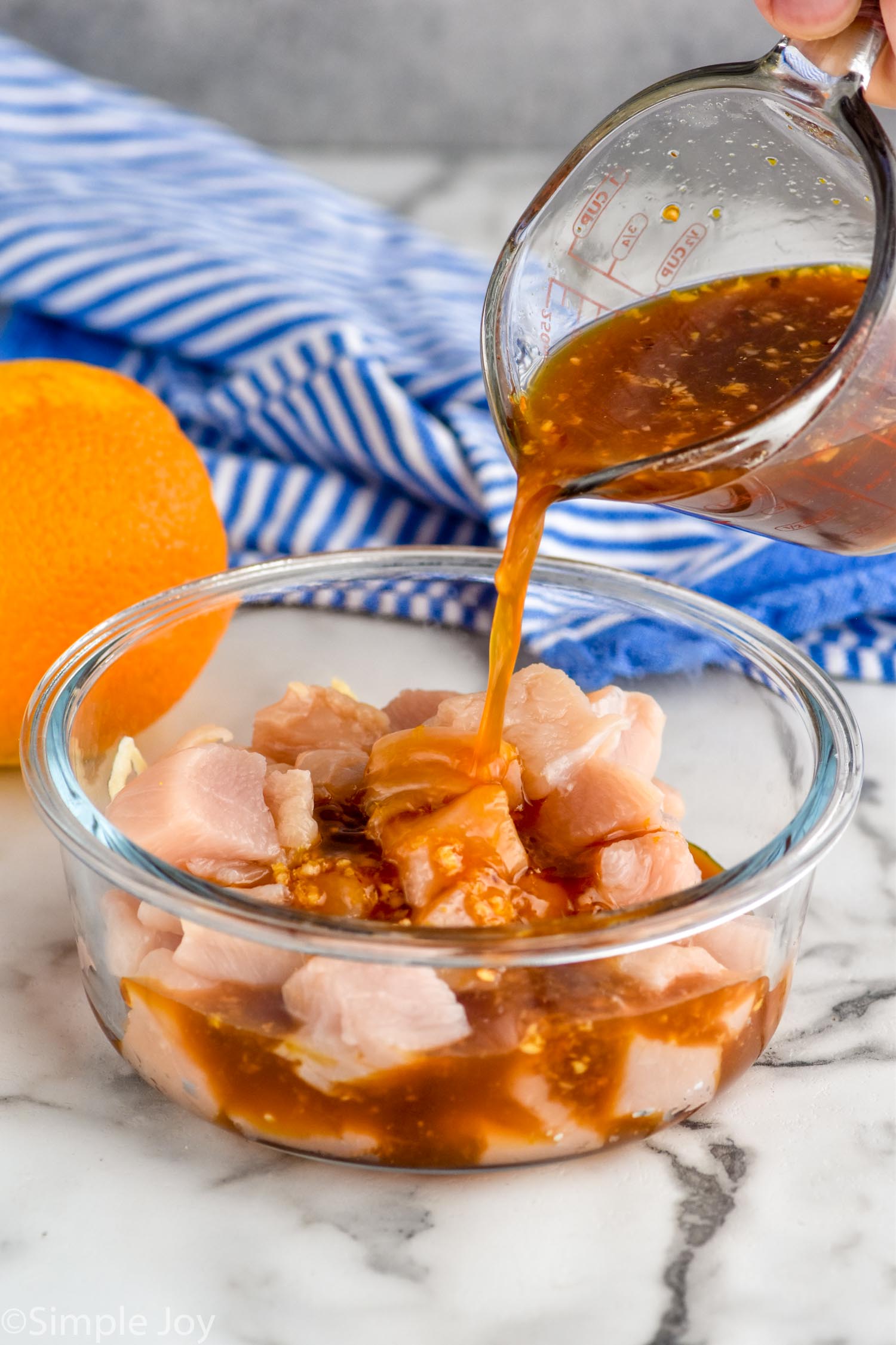 Photo of person's hand pouring measuring cup of ingredients into a container with raw chicken pieces for Orange Chicken recipe.