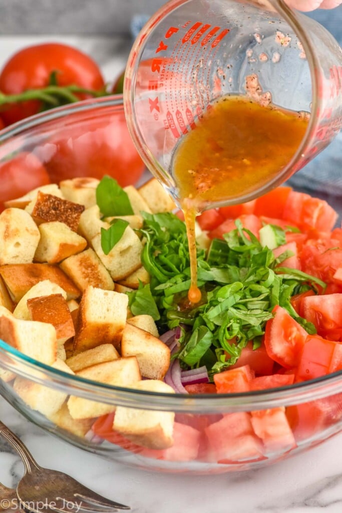 Side view of a glass measuring cup pouring dressing over a bowl of ingredients for Panzanella Salad recipe.