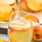 side view of glass of Peach Sangria Recipe garnished with peach slice. More glasses in the background, Extra peaches beside glasses.
