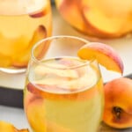 side view of glass of Peach Sangria Recipe garnished with peach slice. More glasses in the background, Extra peaches beside glasses. - 11