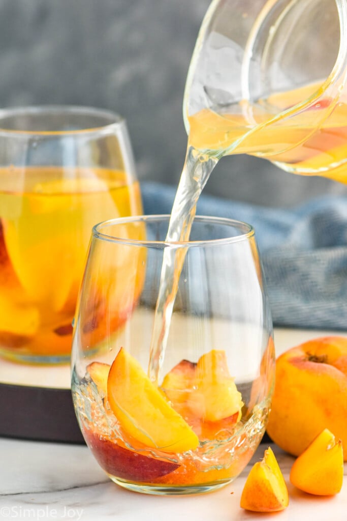 Photo of pitcher of Peach Sangria Recipe pouring into glass of peaches. Extra peaches beside glass.