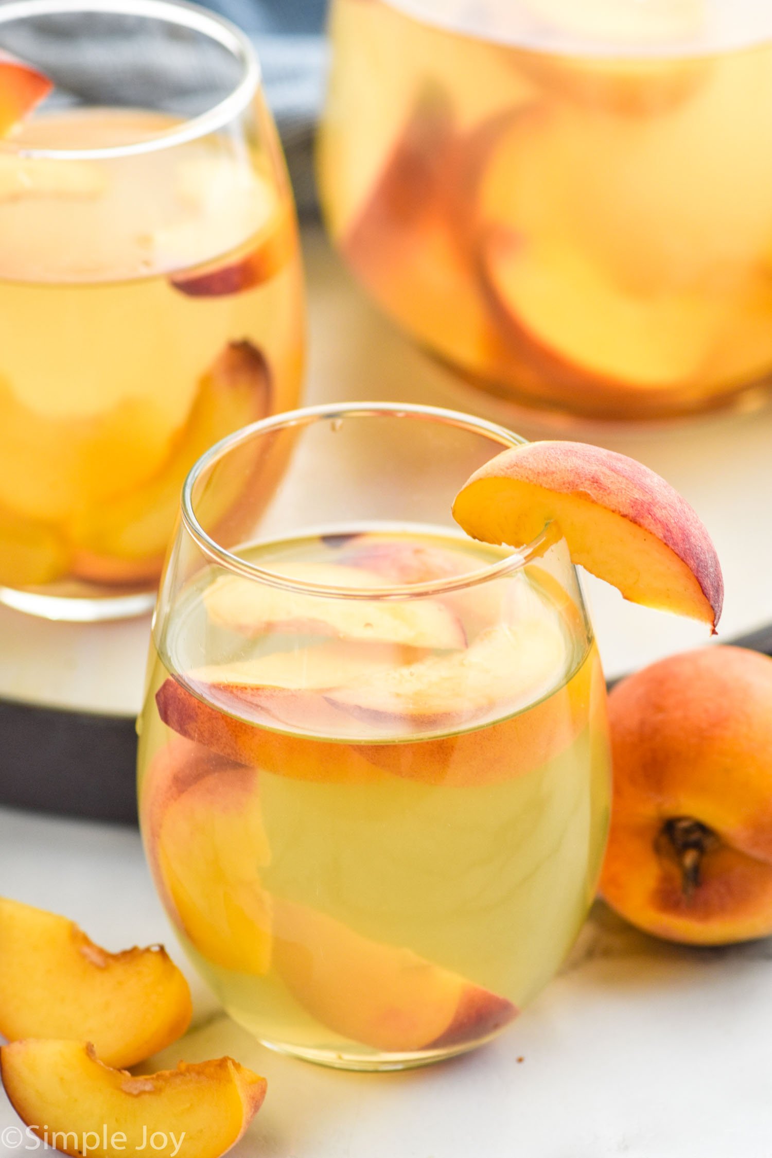 side view of glass of Peach Sangria Recipe garnished with peach slice. More glasses in the background, Extra peaches beside glasses.