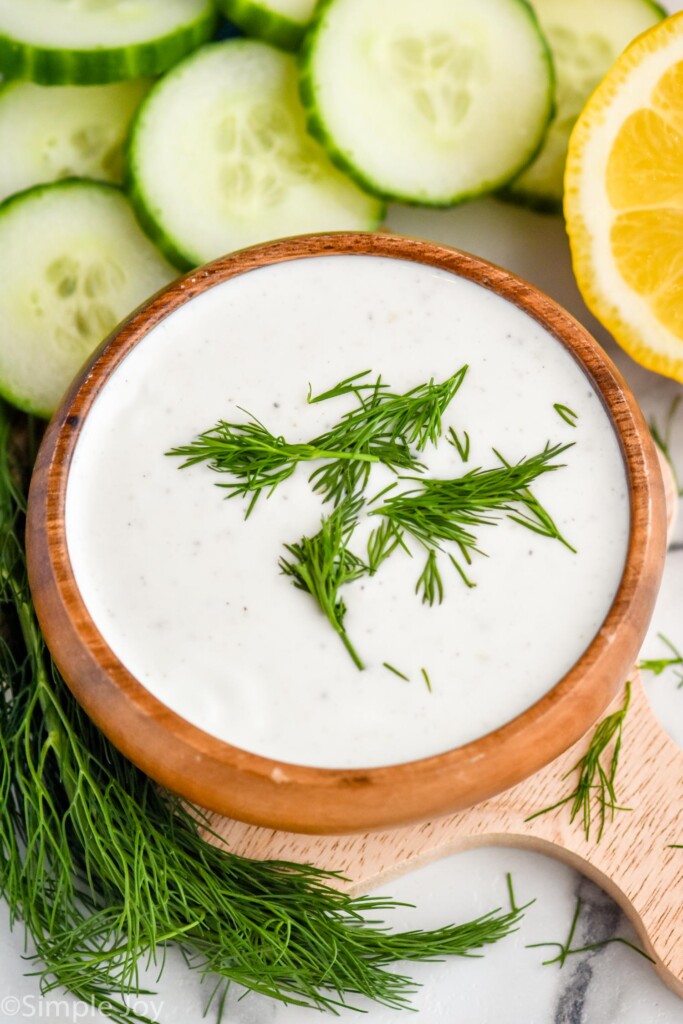 Overhead photo of a bowl of Yogurt Sauce garnished with dill. Dill, cucumber slices, and lemons sit beside bowl.