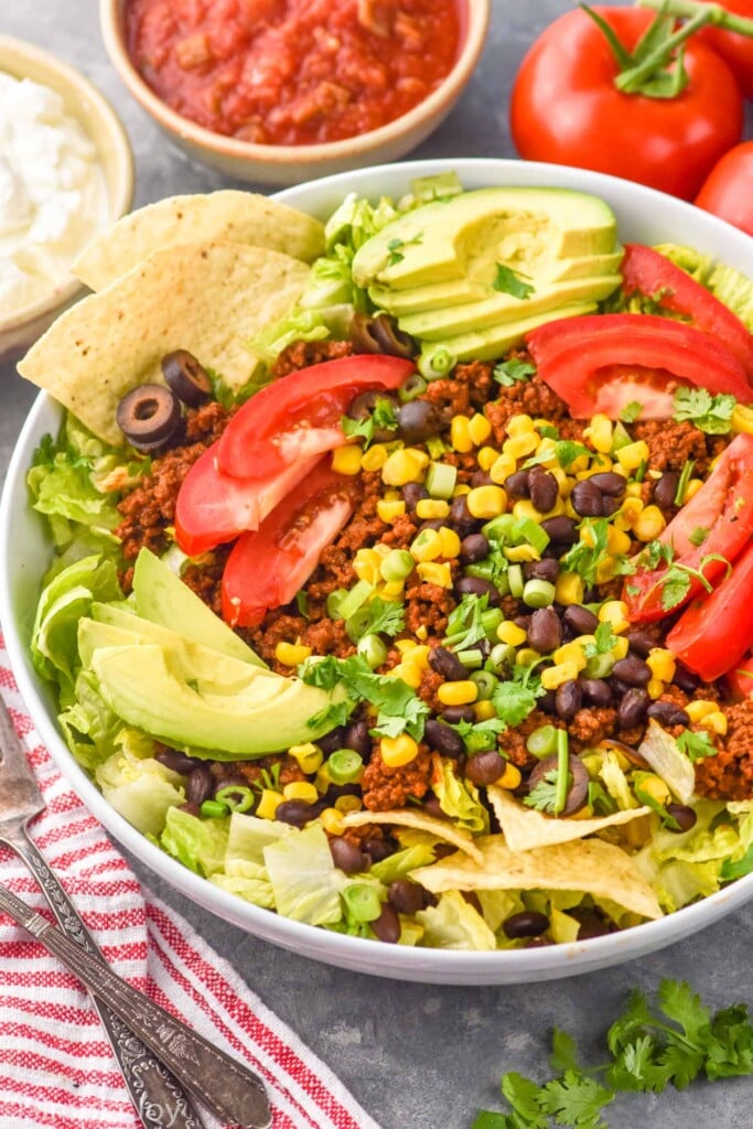 Overhead photo of Taco Salad served in a bowl. Tomatoes, forks, salsa, and sour cream beside bowl.