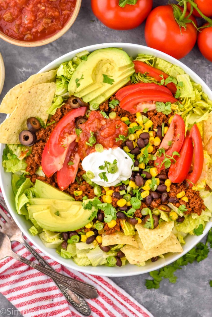 Overhead photo of Taco Salad served in a bowl next to bowl of salsa, extra tomatoes, and forks for eating.