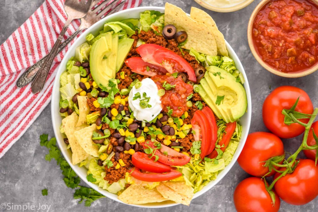 Overhead photo of Taco Salad served in a bowl next to bowl of salsa, extra tomatoes, and forks for eating.