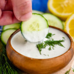 Close up photo of person's hand dipping a cucumber slice into a bowl of Yogurt Sauce. Dill, cucumber slices, and lemons sit beside bowl.