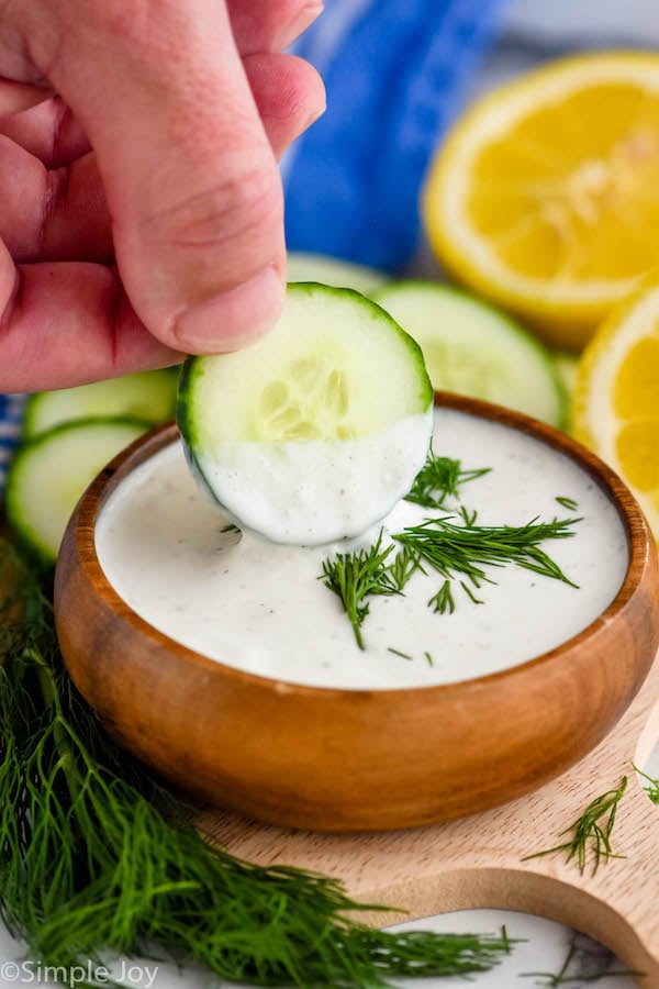 Close up photo of person's hand dipping a cucumber slice into a bowl of Yogurt Sauce. Dill, cucumber slices, and lemons sit beside bowl.