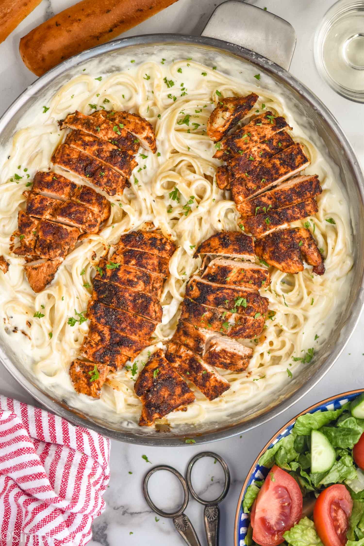 overhead view of a pan of Blackened Chicken Alfredo with breadsticks, bowl of salad, tongs, and towel sitting beside.