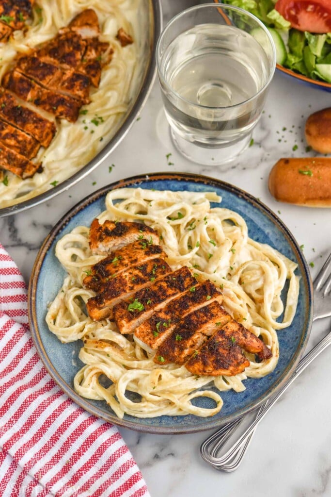 plate of Blackened Chicken Alfredo with pan of Blackened Chicken Alfredo, bowl of salad, breadsticks, and glass of water sitting beside.