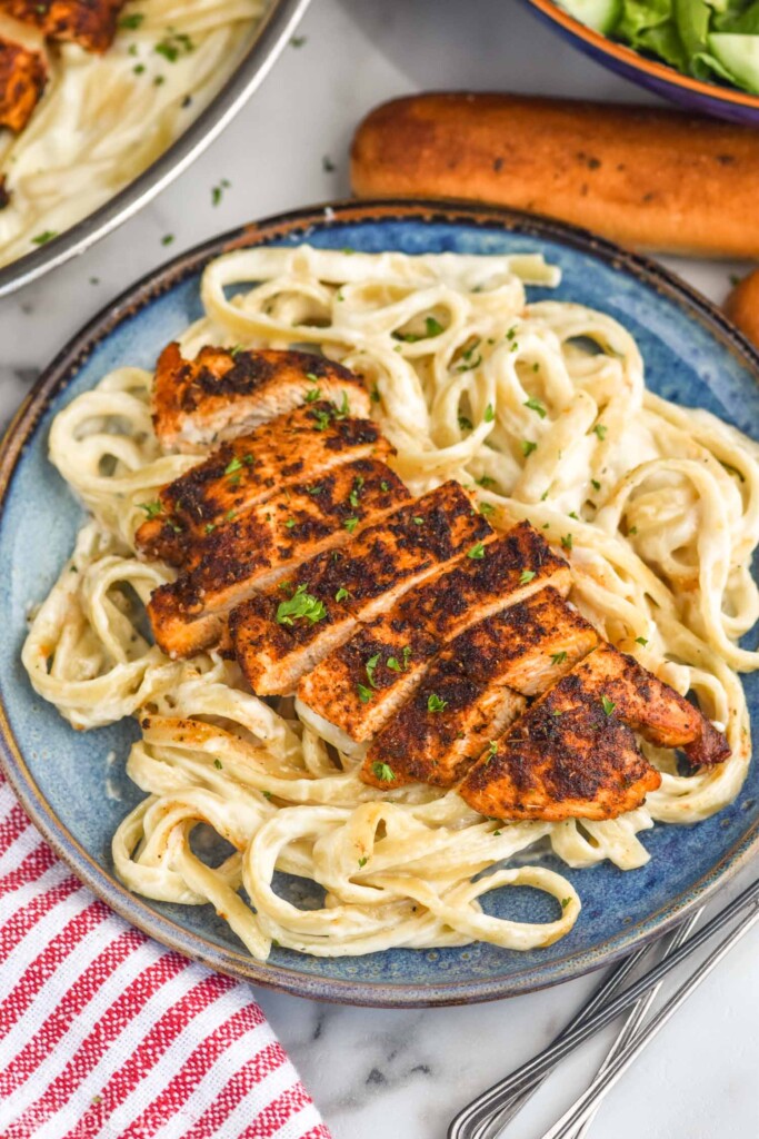 plate of blackened chicken fettuccine alfredo with breadstick and bowl of salad in background