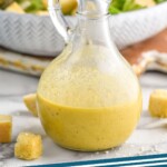Pinterest graphic for Caesar Dressing. Image shows a container of homemade caesar dressing. Bowl of caesar salad sitting in background, croutons sitting beside. Text says "Caesar Dressing simplejoy.com"