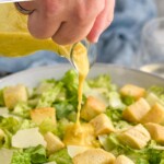 Pinterest graphic for Caesar Dressing Recipe. Text says "the best caesar dressing simplejoy.com" Image shows man's hand pouring container of Caesar Dressing onto a caesar salad with croutons and parmesan cheese.