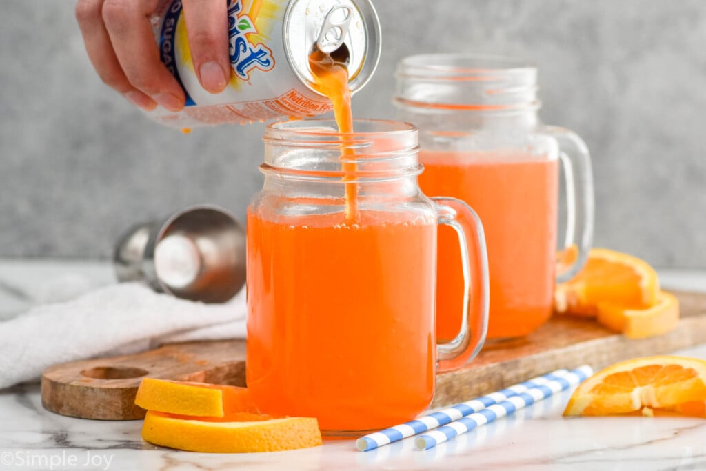 Side view of person's hand pouring orange soda into glass mug for Orange Creamsicle recipe. Another mug of ingredients, orange slices for garnish, and straws on the counter. Cocktail jigger in the background.