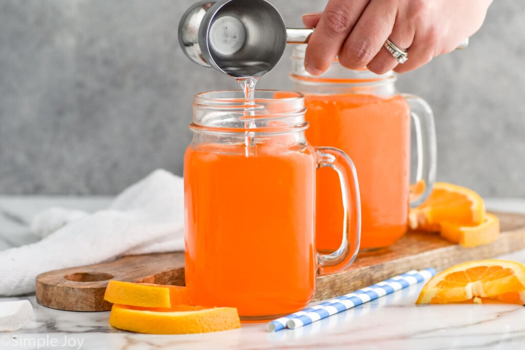 Side view of person's hand pouring cocktail jigger of vodka into mug of orange soda for Orange Creamsicle recipe. Another mug of ingredients, orange slices, and straws on counter.
