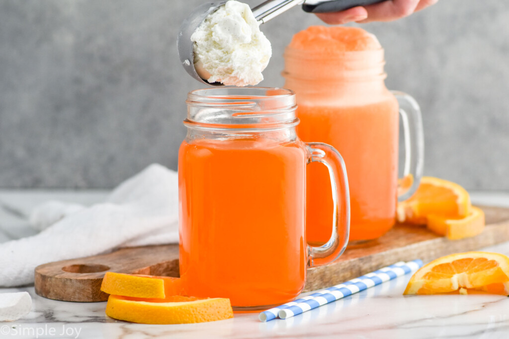 Side view of person's hand holding scoop of ice cream over mug of ingredients for Orange Creamsicle recipe. An Orange Creamsicle, orange slices, and straws on counter.