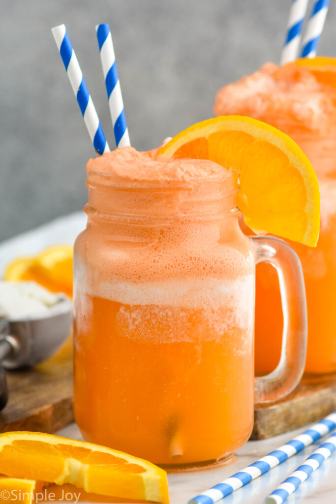 Side view of Orange Creamsicle served in a glass mug garnished with orange slice and straws. Another Orange Creamsicle, orange slices, and straws on counter.
