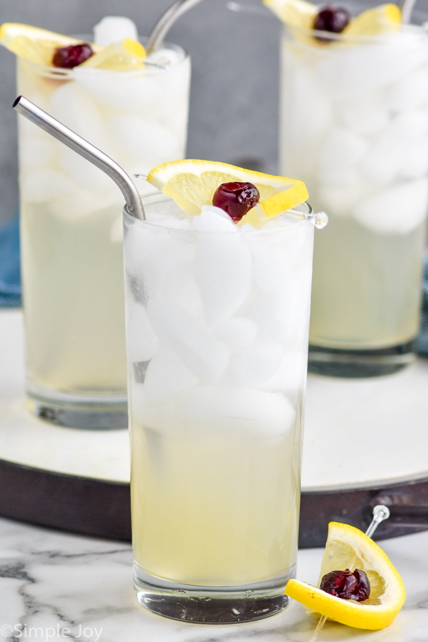 Three glasses of Tom Collins cocktails with ice and garnished with a lemon wedge and cherry.