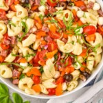 Pinterest graphic for Tortellini Pasta Salad recipe. Image is overhead photo of a bowl of Tortellini Pasta Salad. Serving spoons and tomatoes beside bowl. Text says, "super easy Tortellini Pasta Salad simplejoy.com"