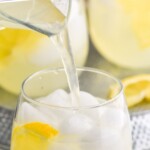 Pinterest graphic for Vodka Lemonade recipe. Text says, "the best Vodka Lemonade simplejoy.com." Image is side view of pitcher of liquid being poured into glass of ice and lemon slice for Vodka Lemonade recipe. More glasses and extra lemon wedges beside glass.