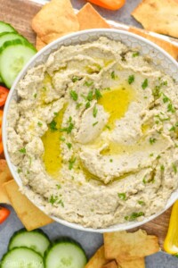 Overhead view of a bowl of Baba Ganoush surrounded by slices of cucumber and pepper and pita chips for serving