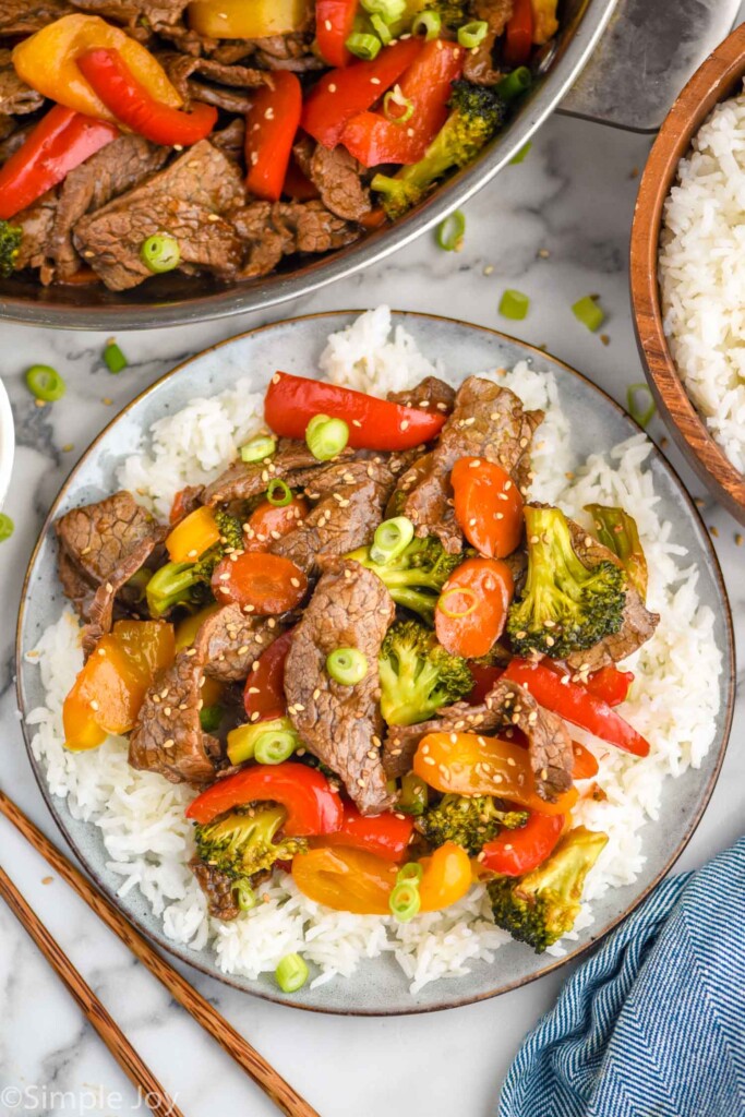 Overhead view of plate of beef stir fry over white rice. Pan of beef stir fry, bowl of white rice, and chopsticks sitting beside.