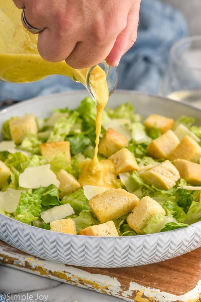 man's hand pouring caesar dressing onto a caesar salad topped with croutons and freshly grate parmesan cheese
