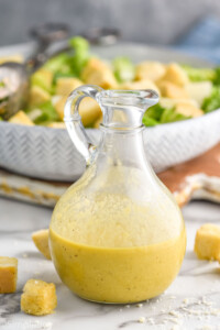 container of homemade Caesar dressing recipe. Bowl of caesar salad and croutons sitting in background