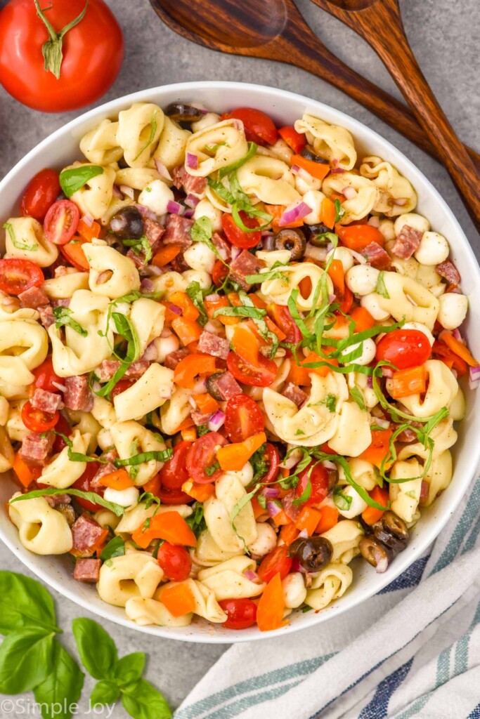 Overhead photo of a bowl of Tortellini Pasta Salad. Serving spoons and tomato beside bowl.