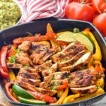Grill pan of chicken fajitas with lime wedges for serving. Bowl of guacamole and fresh tomatoes sit in background