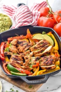 Grill pan of chicken fajitas with lime wedges for serving. Bowl of guacamole and fresh tomatoes sit in background